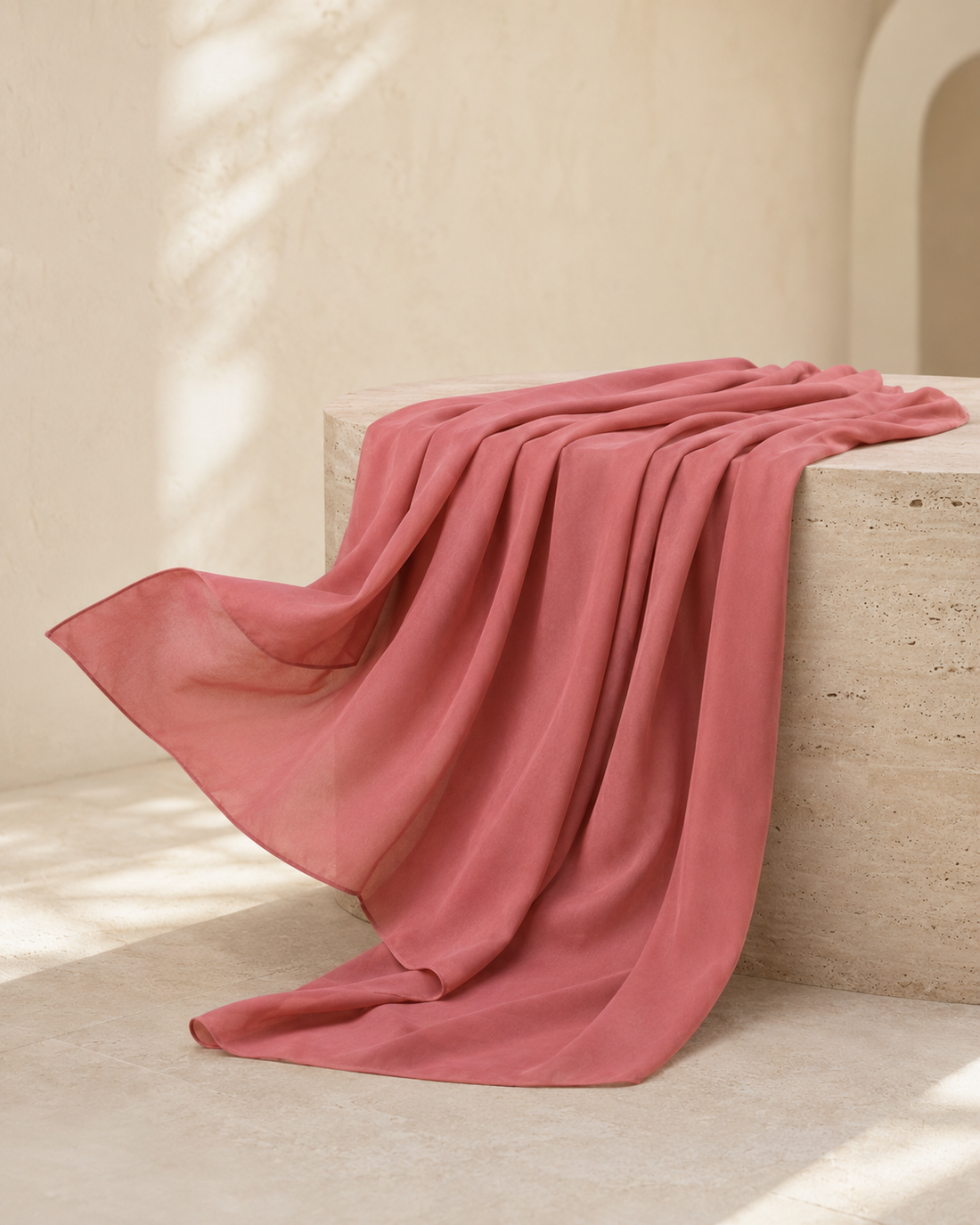 A dusty rose pink lightweight scarf draped elegantly over a textured stone block in a warm, sunlit minimalist interior.